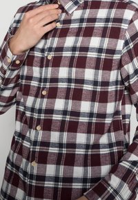 Man som knäpper den övre knappen på en långärmad murrich, vit och svart rutig skjorta med beige knappar mot en neutral bakgrund.