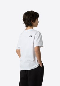 Bela bombažna majica z okroglim izrezom in kratkimi rokavi, z majhnim črnim logotipom na zgornjem delu hrbta. Enostaven dizajn, sproščen kroj.