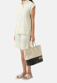 Camicetta senza maniche di colore giallo chiaro con colletto, abbinata a pantaloni corti leggeri. Borsa a tracolla beige e nera con logo "BOGNER". Sandali bianchi con cinturini.