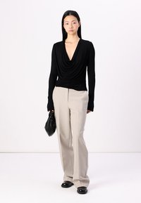 Black long-sleeve top with a draped neckline, paired with beige wide-leg pants. The model holds a black handbag. Smooth texture and fitted design.