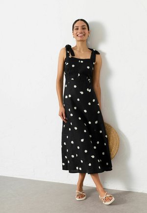 Woman smiling wearing a black sleeveless midi dress with white polka dots, holding a straw bag, and white strappy sandals, standing indoors.