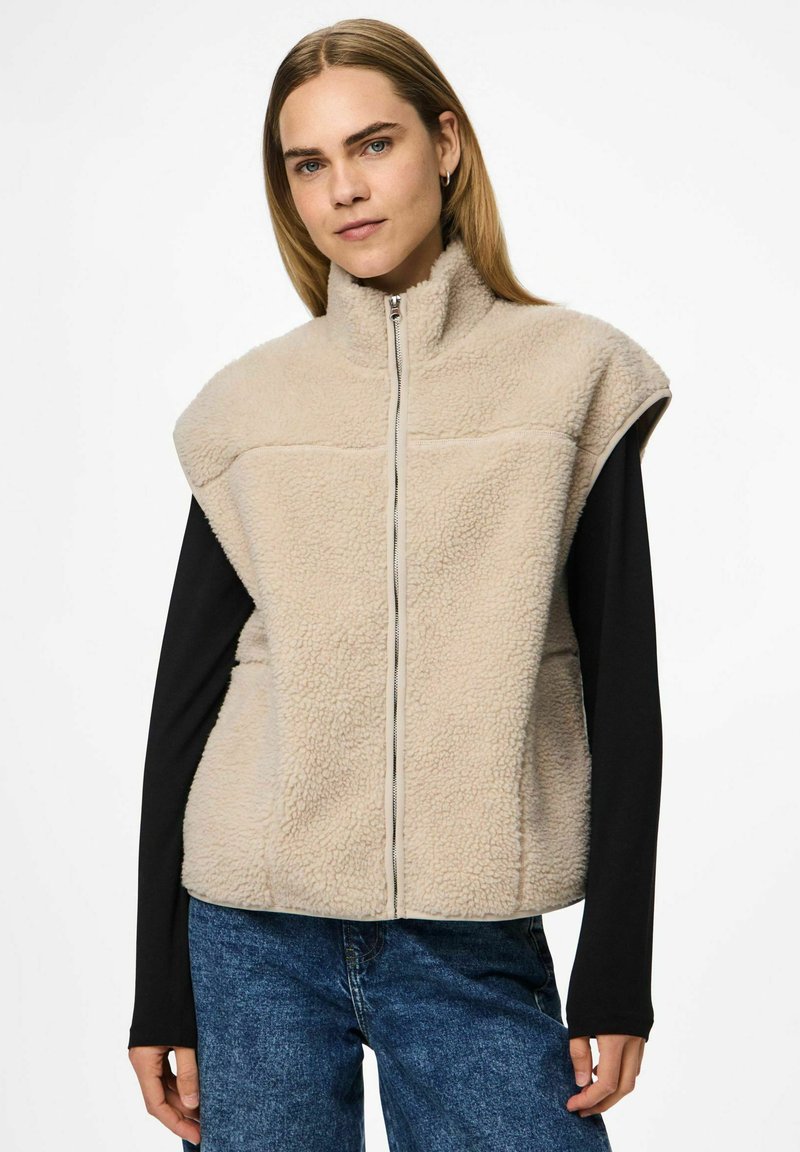 Vrouw in een beige fleece zip-up vest over een zwart shirt met lange mouwen en blauwe jeans, staand tegen een effen achtergrond.
