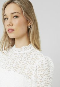 Blouse en dentelle blanche avec un col montant et des manches bouffantes, présentant des motifs floraux complexes et une texture transparente. Des boucles d'oreilles en argent complètent le look.