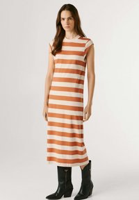 Žena stojaca v bezrukávových šatách po kolená s oranžovými a krémovými horizontálnymi pruhmi a čiernymi čižmami po lýtka na jednoduchej pozadí.