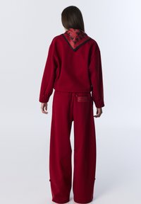 Sudadera roja de gran tamaño con mangas largas, que presenta un cuello tipo bufanda con un patrón rojo acentuado en negro. Pantalones rojos holgados con botones de presión en los laterales.