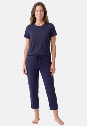Femme debout, pieds nus, portant un t-shirt bleu marine à manches courtes et un pantalon court à cordon sur un fond clair uni.