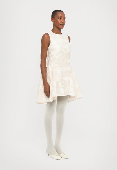 Beige mouwloos jurkje met een uitlopende rok, textuurstof en een subtiel patroon. Gecombineerd met lichtgrijze panty’s en crème hakken.