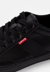 Baskets Levi's noires avec lacets noirs, panneaux en cuir et tissu, et une petite étiquette logo Levi's rouge près des œillets.
