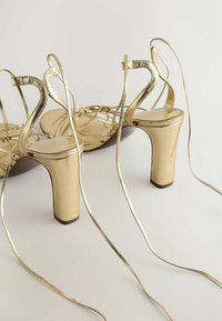 Sandales à talons hauts dorées avec des brides à la cheville et de longs lacets fins s'étendant vers l'extérieur sur un fond clair.