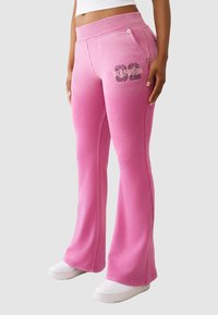 Różowe bawełniane spodnie dresowe o rozszerzanym kroju, z nadrukiem logo i boczną kieszenią zapinaną na zatrzask. Gładka tekstura i elastyczna talia.
