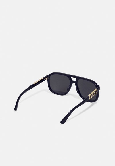 Lunettes de soleil noires avec des verres sombres, monture rectangulaire et accents dorés sur les branches, arborant le logo. Texture en plastique lisse.