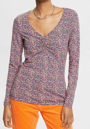 Femme portant un haut à manches longues, floral multicolore, avec un devant froncé et un pantalon en velours côtelé orange vif.
