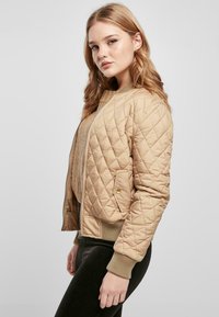 Beige quilted bomberjakke med rund hals, ribbestrikkede mansjetter og glidelås. Har sidelommer og en glatt tekstur.