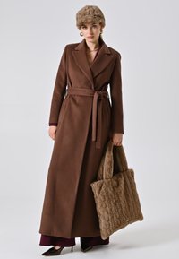Donna con lungo cappotto marrone con cintura, berretto di pelliccia marrone, che tiene una grande borsa di pelliccia texturizzata marrone, indossa orecchini d'oro e tacchi a punta neri.