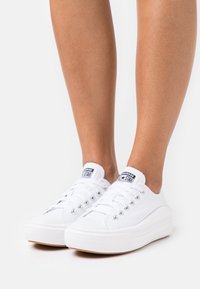 Baskets en toile blanche avec un design plat, un bout rond et une semelle en caoutchouc texturée. Elles présentent des œillets argentés et une petite étiquette sur la languette.