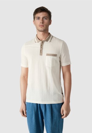 Ung mand iført en cremefarvet kortærmet poloshirt med beige krave og lomme, kombineret med blå bukser, stående foran en ensfarvet baggrund.