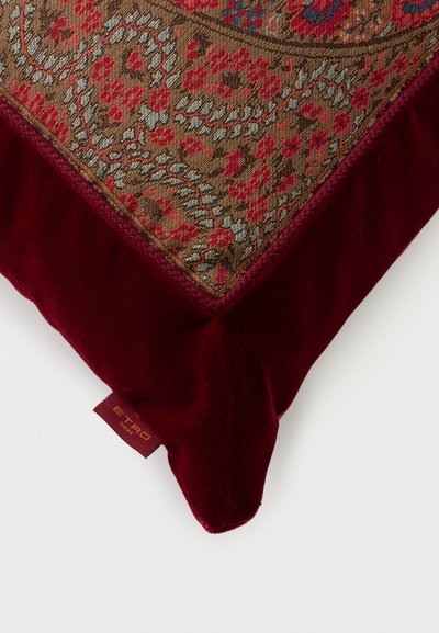 Coussin carré avec des bords en velours rouge foncé et un centre en tapisserie florale complexe aux tons rouges, bleus et bruns, avec une petite étiquette de marque en tissu.