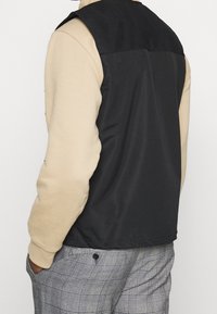 Black and tan sleeveless jacket with a smooth, shiny finish; paired with gray plaid pants featuring a slight texture and a button detail.