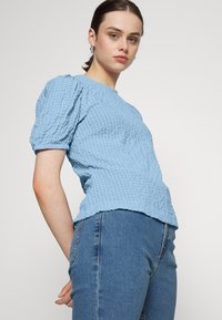 Blusa azul clara texturizada com mangas curtas bufantes, decote redondo e barra desigual. Combinada com jeans justos azul escuro.