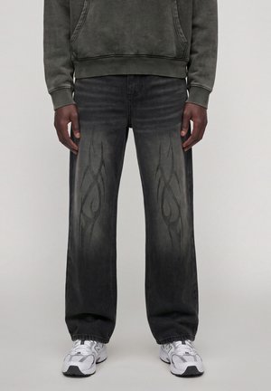 Personne portant un jean gris foncé délavé avec des motifs tribaux noirs, un sweat-shirt noir-vert délavé et des baskets blanches, debout droit.