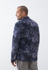 Chaqueta de cordón en tintes anudados en azul marino y gris, con rayas verticales, cuello y bolsillos laterales, llevada por un hombre, mostrando un patrón texturizado.