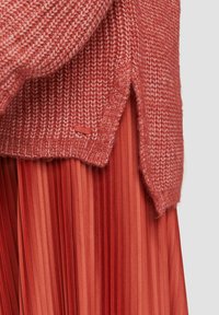 Dzianinowy sweter w miękkim, teksturowanym kolorze koralowym z długością wysoka-niska i bocznymi rozcięciami, zestawiony z plisowaną spódnicą koralową w warstwowych odcieniach.