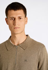 Brun zip-up tröja med platt krage, strukturerad tyg och liten logotyp på vänster sida. Modellen har övre delen av kroppen synlig mot en ljus bakgrund.