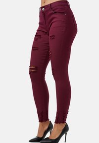 Jeans skinny burdeos con detalles desgastados y dobladillos deshilachados, hechos de material elástico. Combinados con tacones negros de punta para un contraste.