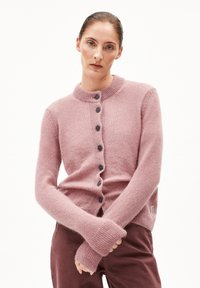 Frau mit nach hinten gegeltem Haar, die einen staubigen rosa, geknöpften Cardigan und maroonfarbene Hosen trägt, mit verschränkten Armen vor einem schlichten weißen Hintergrund.