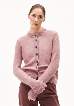 Femme aux cheveux tirés en arrière, portant un cardigan à boutons rose poussiéreux et un pantalon bordeaux, les bras croisés devant un fond blanc uni.