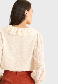 Koronkowa bluzka w kolorze off-white z falbaniastym kołnierzem i długimi puffowanymi rękawami. Całość pokryta kwiatowym wzorem. Noszona z brązowymi spodniami z sztruksu.