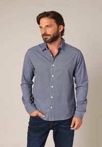 Donkerblauw overhemd met knopen, met een klein wit patroon, lange mouwen en een kraag. Gekozen met donkere spijkerbroek, handen in de zakken.