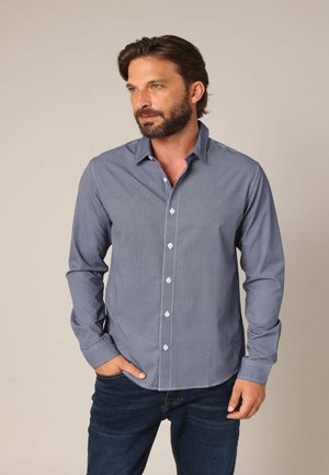 Chemise boutonnée bleu marine avec un petit motif blanc, manches longues et col. Portée avec un jean en denim foncé, les mains dans les poches.