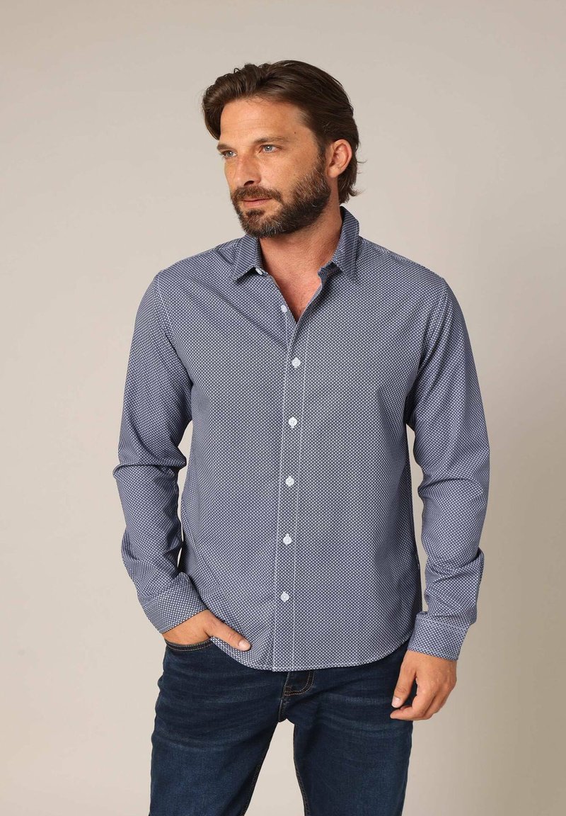 Donkerblauw overhemd met knopen, met een klein wit patroon, lange mouwen en een kraag. Gekozen met donkere spijkerbroek, handen in de zakken.