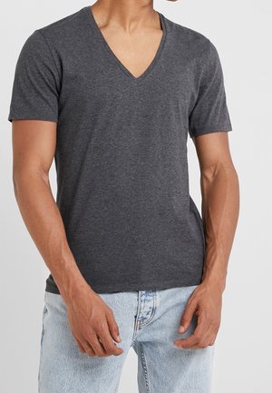 Homme portant un T-shirt à col en V gris foncé à manches courtes et un jean en denim bleu clair, se tenant avec les mains détendues le long des hanches.