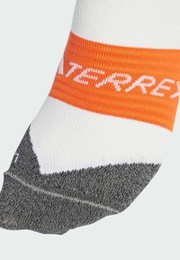 Biela a sivá ponožka s oranžovým pruhovaným pásom, na ktorom je bielym písmom vytlačené slovo "TERREX". Výrazný dizajn s textúrovanou tkaninou v priliehavom tvare.