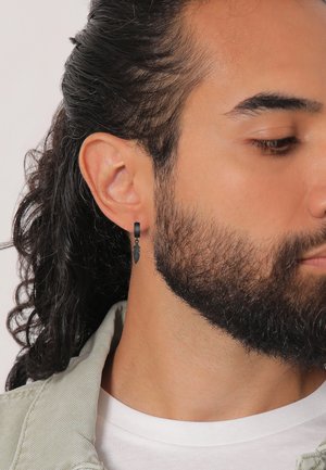 Boucle d'oreille en plumes noires avec un anneau circulaire lisse, porté à l'oreille gauche d'une personne avec des cheveux foncés de longueur moyenne et une barbe.