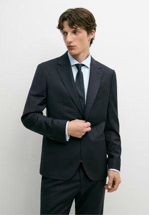 Giovane uomo che indossa un abito blu scuro, camicia azzurra e cravatta scura, sistema la giacca su uno sfondo bianco semplice.