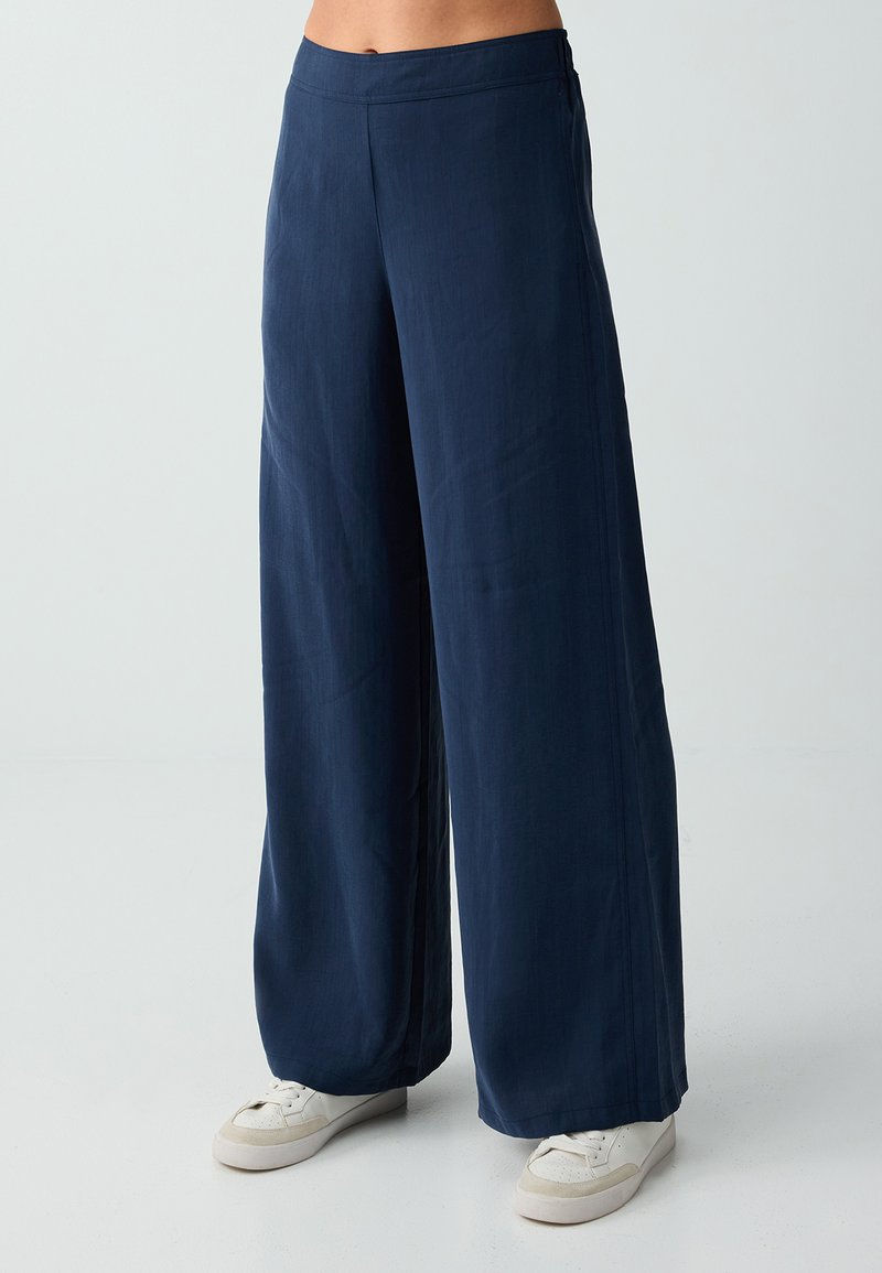 Personne portant un pantalon large bleu marine et des baskets blanches, debout devant un fond clair uni.