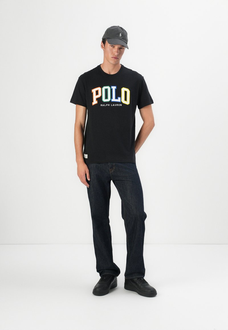 Czarna koszulka z wielokolorowym napisem "POLO" oraz "RALPH LAUREN" pod spodem, w połączeniu z ciemnymi dżinsami i czarnymi sneakersami. Szara czapka na modelu.