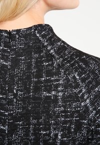 Svart texturerad tyg med ett vitt mönster, med hög krage och dragkedjedetalj bak. Avslappnad design för vardagsbruk.