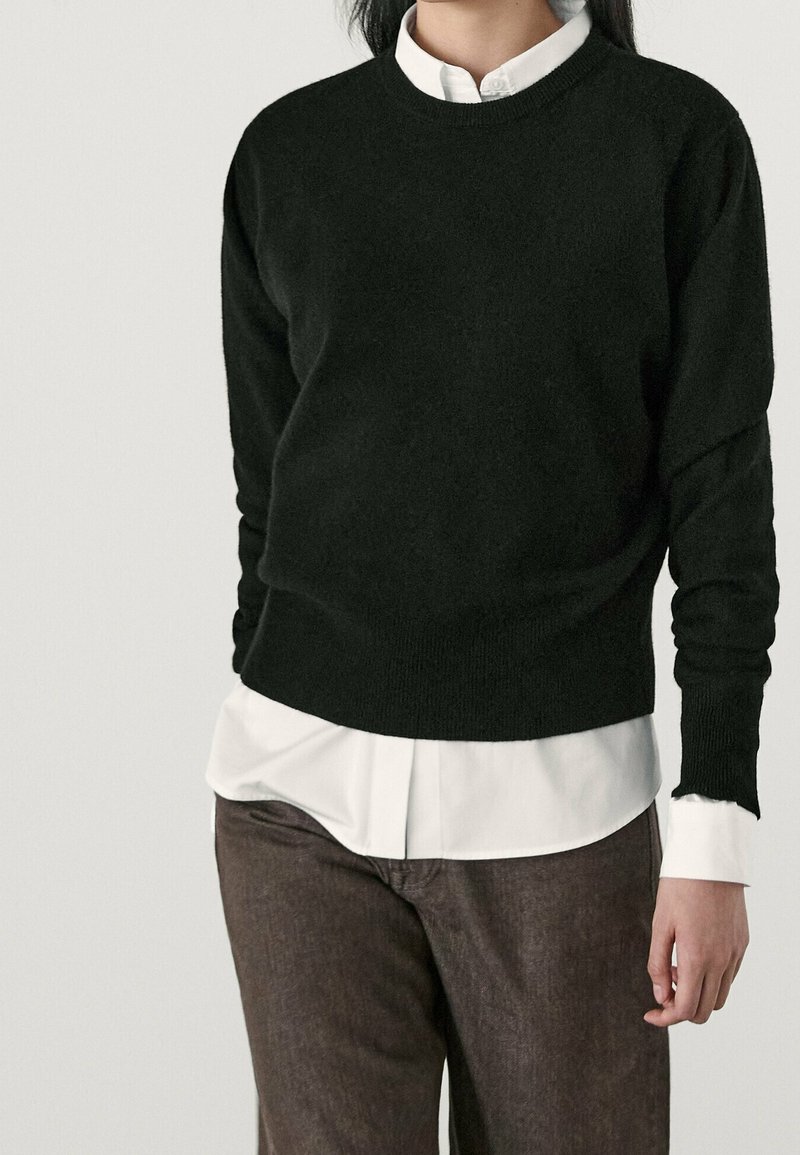 Femme portant un pull noir par-dessus une chemise blanche à col et un pantalon marron, debout devant un fond clair uni.
