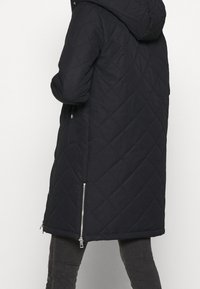 Abrigo negro acolchado con capucha, mangas largas y bolsillos laterales con cremallera, con una longitud a mitad del muslo y tela texturizada.