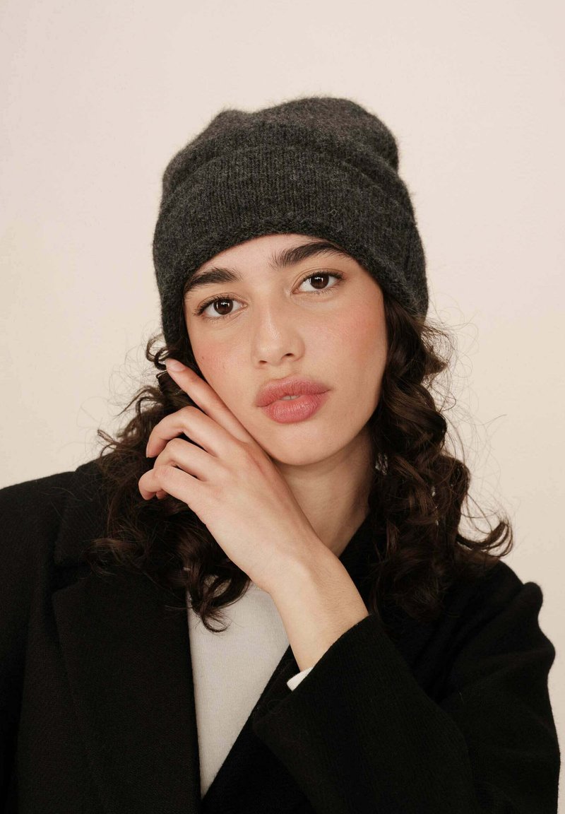 Béret en tricot gris foncé, en texture douce, avec un design replié, associé à un manteau noir et une chemise claire en dessous.