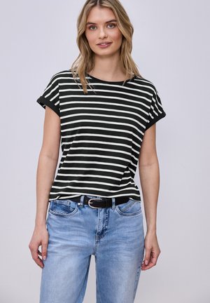 Frau, die ein schwarz-weiß gestreiftes Kurzarmshirt trägt, das in hellblaue Jeans mit einem schwarzen Gürtel gesteckt ist, stehend vor einem einfarbigen Hintergrund.