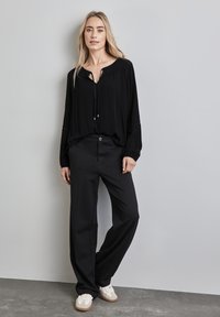 Zwarte lange mouw top met een V-hals en trekkoorddetails, gecombineerd met zwarte wijde broek en lichte sneakers. Eenvoudige, casual look.
