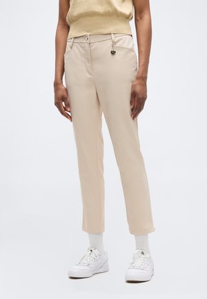 Person iført beige bukser med lige ben og en lille sort og guld charm på bæltestrop, kombineret med hvide sokker og hvide sneakers.