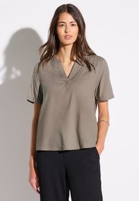 Blusa elegante a maniche corte in verde oliva, con scollatura a V, texture liscia e vestibilità rilassata. Abbinata a pantaloni neri con texture.