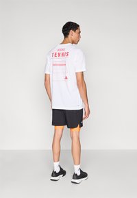 Fehér Adidas póló, amelyen piros "TENNIS" felirat és pályagrafika található a hátoldalon. Fekete rövidnadrággal és fekete cipővel párosítva.