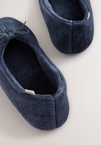 Paire de chaussons bleu marine en velours côtelé avec petit nœud sur le dessus, intérieur doux matelassé et étiquette de marque en tissu à l’intérieur.